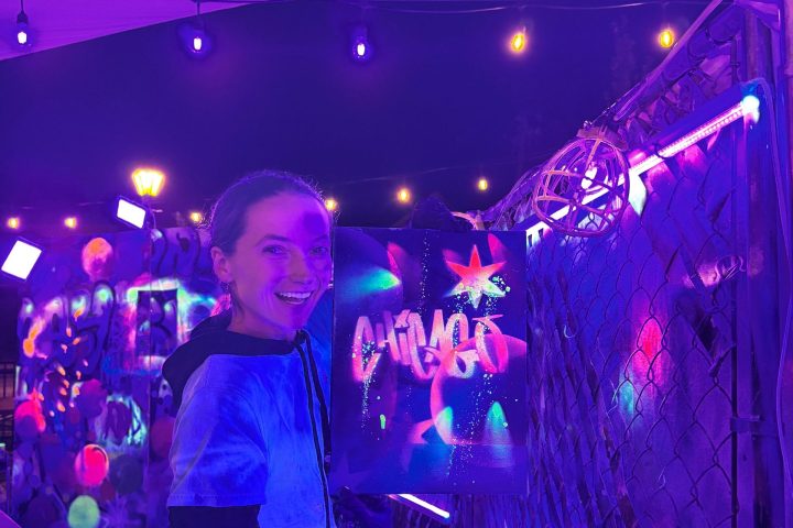 Person under purple lights holding neon spray paint art with the word 'Chicago' at a night event.